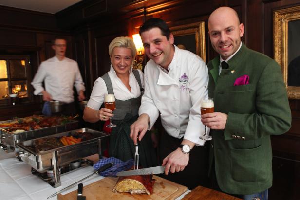 Eine Frau im Dirndl, ein Koch und ein Mann in Tracht stehen an einem Buffet mit bayerischen Spezialitäten und Bier.
