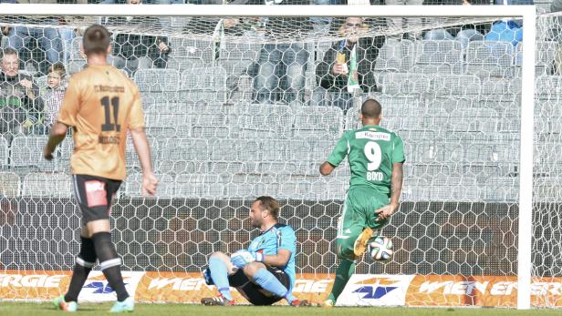 Ein Fußballspieler in Grün schießt den Ball aufs Tor, während der Torwart sitzt.