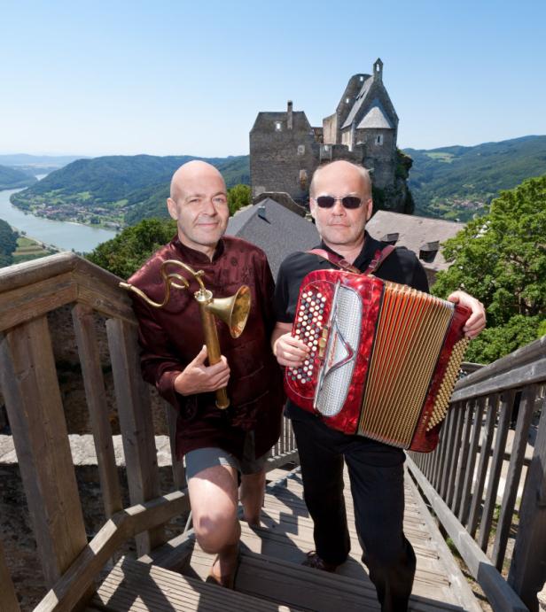 Zwei Männer mit Musikinstrumenten stehen auf einer Holztreppe vor einer Burgruine.