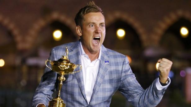 Ein Mann im karierten Sakko jubelt mit einer goldenen Trophäe in der Hand.