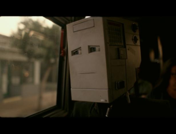 Eine Person mit einem alten Computer als Kopf sitzt in einem Fahrzeug.