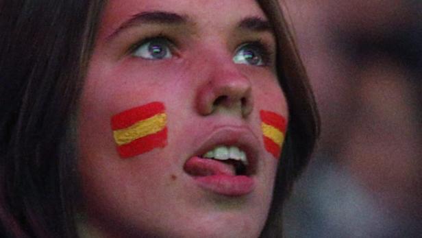 Eine junge Frau mit aufgemalten spanischen Flaggen auf den Wangen schaut erwartungsvoll.