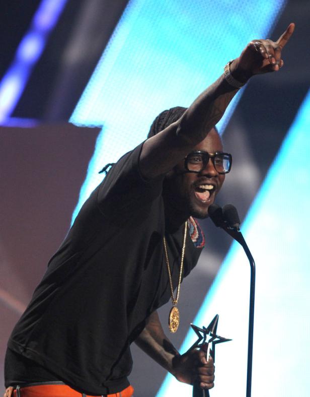 T-Pain hält eine Trophäe in der Hand und gestikuliert auf einer Bühne.