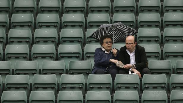 Ein älteres Paar sitzt unter einem gepunkteten Regenschirm auf den grünen Sitzen eines Stadions.