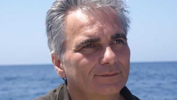 Ein Mann mit grauen Haaren blickt vor einem blauen Meer in die Ferne.