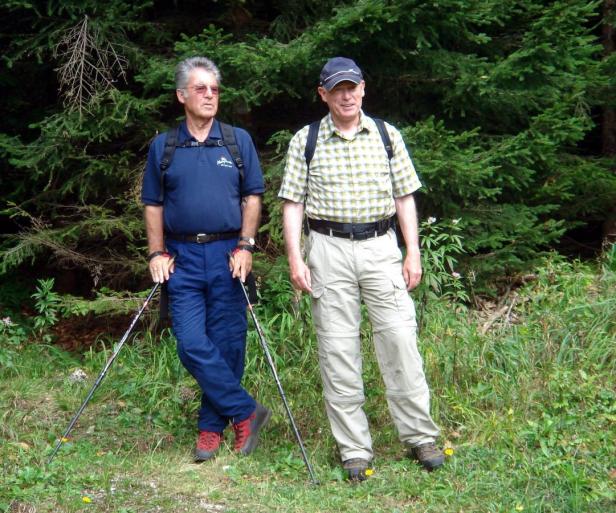 Zwei ältere Männer mit Wanderstöcken stehen vor einem grünen Wald.