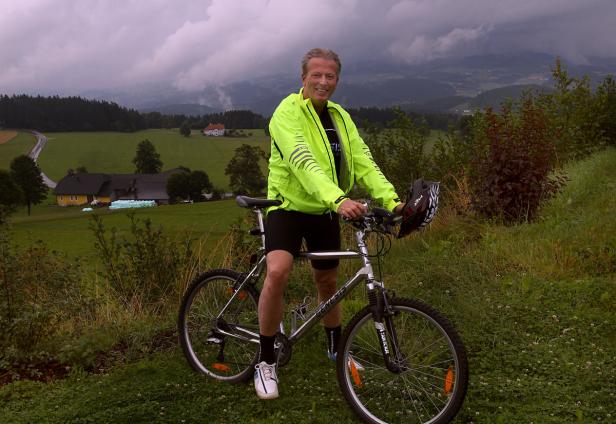 Ein Mann mit leuchtend gelber Jacke posiert mit seinem Fahrrad in einer ländlichen Gegend.