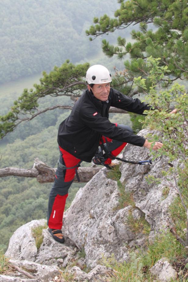 Ein Mann mit Helm klettert an einem Felsen mit Blick auf eine bewaldete Landschaft.