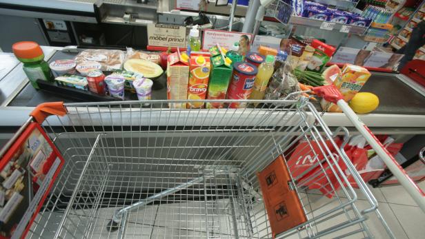 Ein Einkaufswagen steht an der Kasse mit Lebensmitteln wie Joghurt, Saft und Gemüse.
