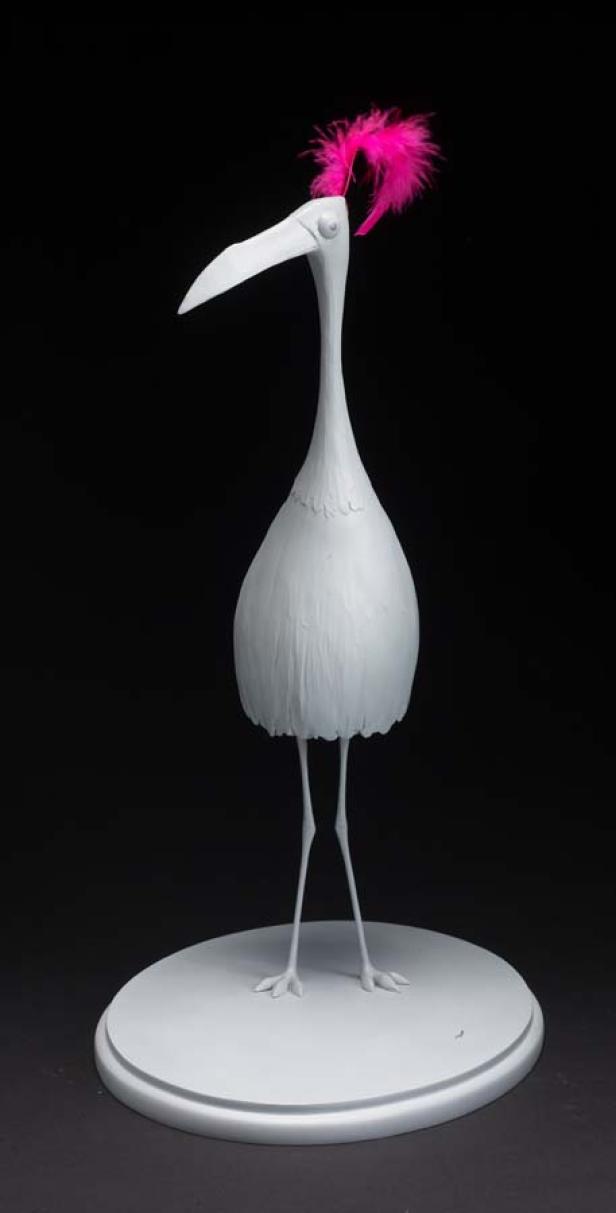 Eine weiße Vogelstatue mit einer pinkfarbenen Feder auf dem Kopf steht auf einem Sockel.