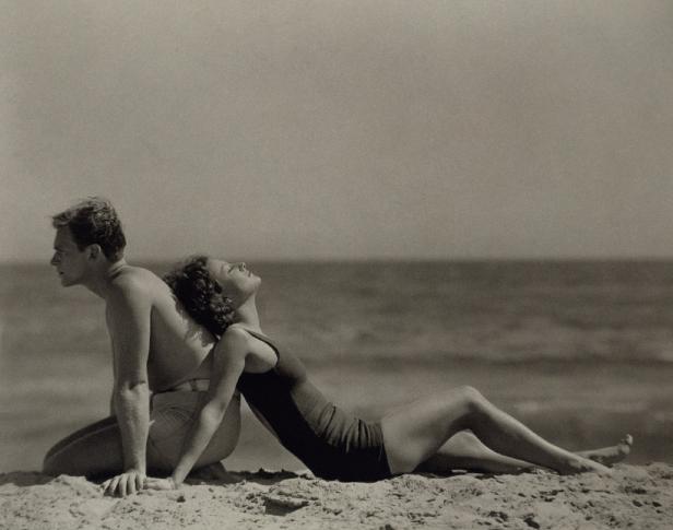 Ein Mann und eine Frau sitzen Rücken an Rücken am Strand.
