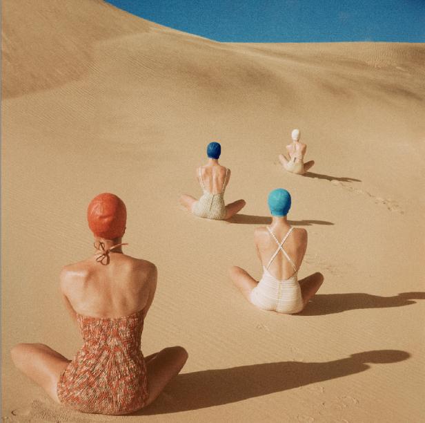 Vier Frauen mit Badebekleidung und Badekappen sitzen in einer Sandwüste.