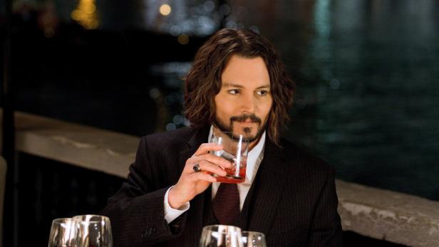 Johnny Depp sitzt an einem Tisch und hält ein Glas mit einem Getränk in der Hand.