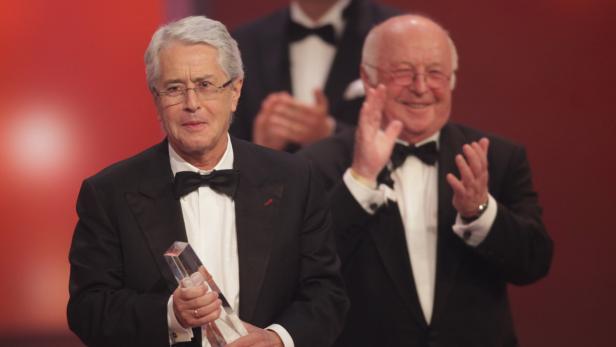 Ein Mann im Smoking hält eine Auszeichnung, während ein anderer Mann applaudiert.