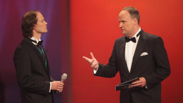 Zwei Männer in Anzügen stehen auf einer Bühne vor einem roten Hintergrund.