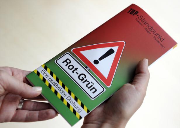 Eine Hand hält eine Broschüre mit einem Warnschild und der Aufschrift „Rot-Grün: Eine gefährliche Drohung“.
