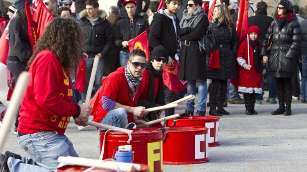 Eine Gruppe von Menschen demonstriert mit roten Fahnen und schlägt auf rote Fässer.