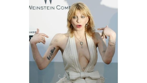 Courtney Love in einem weißen Kleid mit Tätowierungen auf ihren Armen.