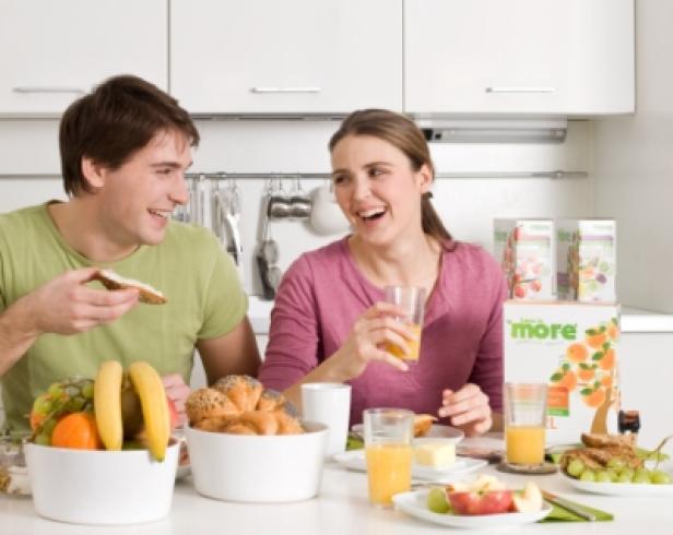 Ein Paar lacht beim Frühstück mit Saft, Brot und Obst in einer hellen Küche.