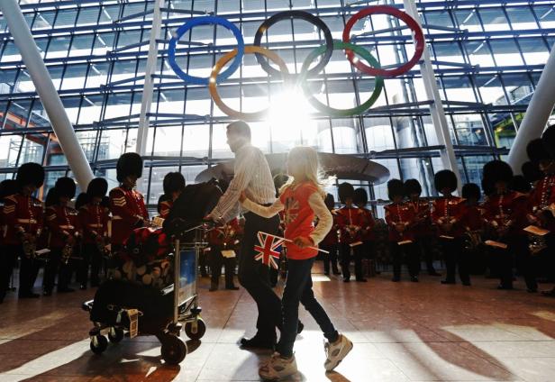 Ein Vater und seine Tochter mit Union-Jack-Flagge gehen an einer Gruppe von Gardesoldaten vorbei; im Hintergrund die olympischen Ringe.