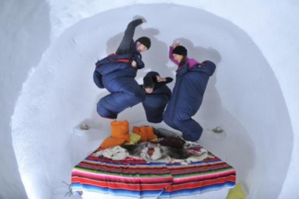 Drei Personen in Schlafsäcken springen über ein Bett in einem Iglu.