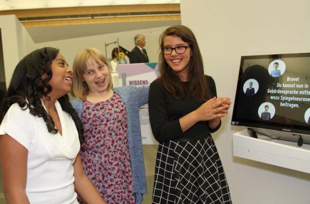Drei junge Frauen stehen lächelnd vor einem Bildschirm mit Gebärdensprache-Informationen.