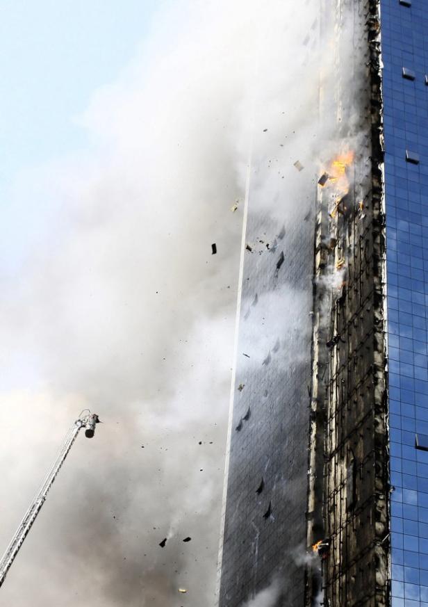 Ein Hochhaus steht in Flammen, Rauch steigt auf und Trümmer fallen herab. Eine Drehleiter ist im Einsatz.