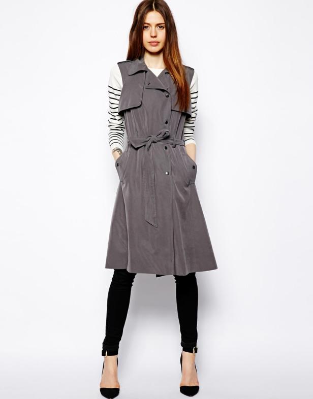 Eine Frau mit braunen Haaren trägt einen grauen, ärmellosen Trenchcoat, schwarze Hose und schwarze Schuhe.