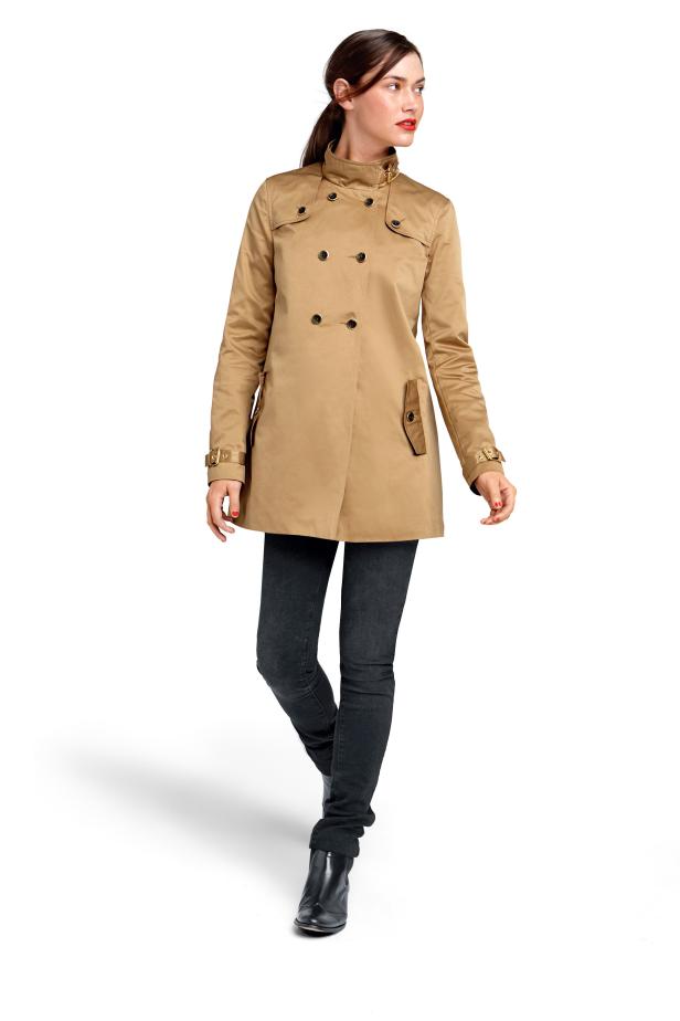 Eine Frau mit braunen Haaren trägt einen beigen Trenchcoat, dunkle Jeans und Stiefeletten.