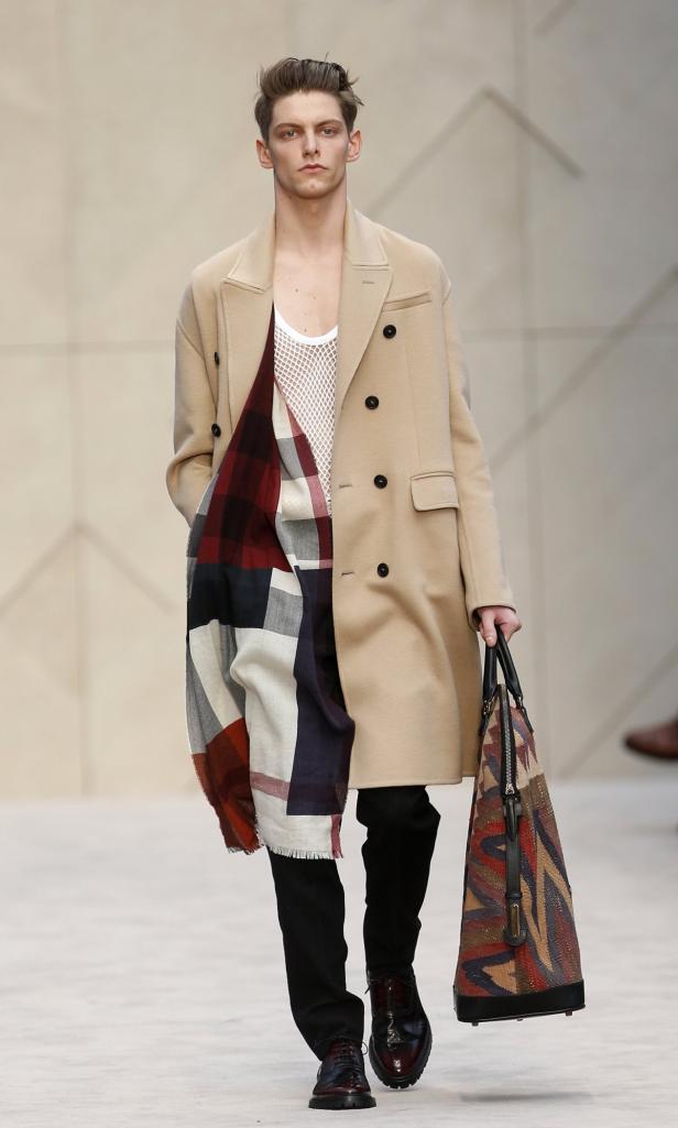 Ein Mann in einem beigen Mantel, einem karierten Schal und einer gemusterten Tasche auf einem Laufsteg.