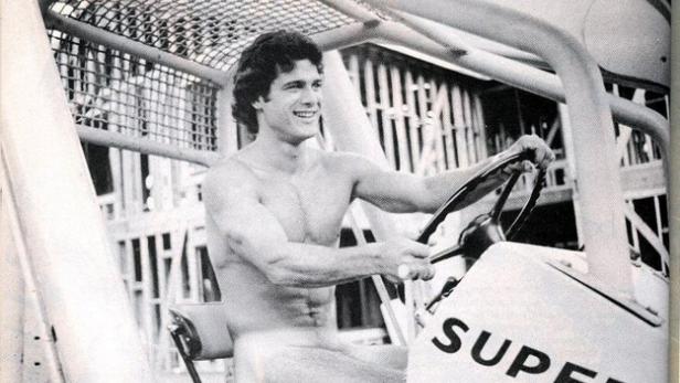 Ein lächelnder, muskulöser Mann fährt ein offenes Fahrzeug mit der Aufschrift „Super“.