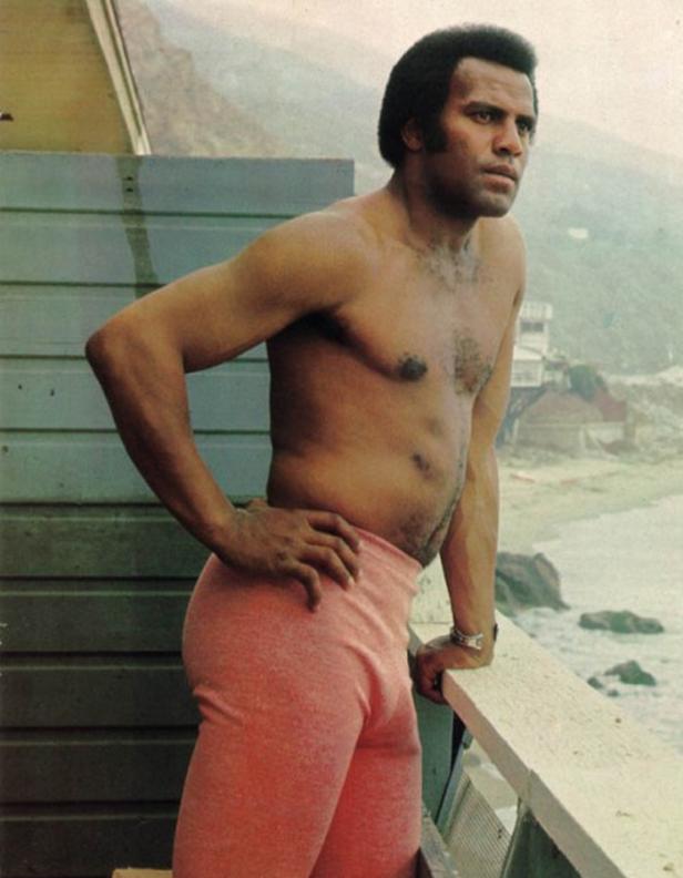 Ein barfußiger Mann in rosafarbener Hose steht auf einem Balkon mit Blick auf das Meer.