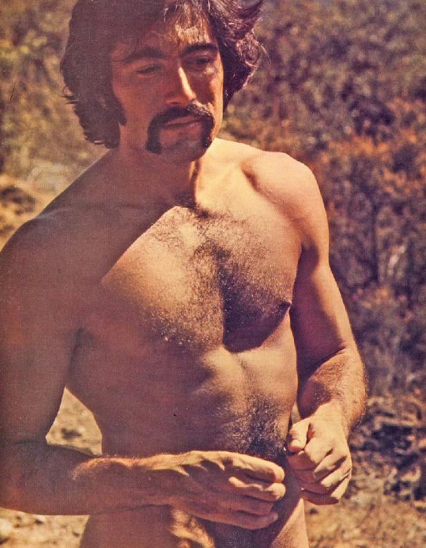 Ein Mann mit Schnurrbart steht barfuß und oberkörperfrei im Freien.