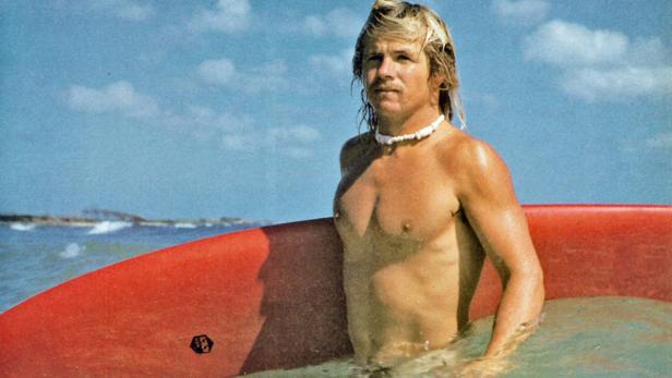 Ein Surfer mit blonden Haaren und einer Muschelkette steht mit seinem Surfbrett im Wasser.