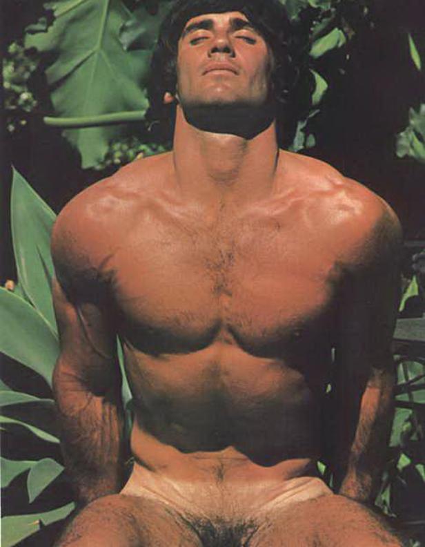 Ein Mann mit freiem Oberkörper sitzt mit geschlossenen Augen vor tropischen Pflanzen.