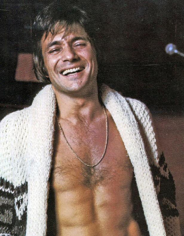 Ein lächelnder Mann mit offenem Strickmantel und Goldkette.