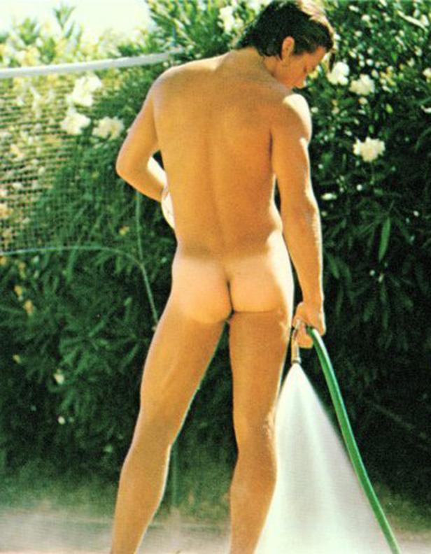 Ein Mann gießt unbekleidet mit einem Gartenschlauch Pflanzen.