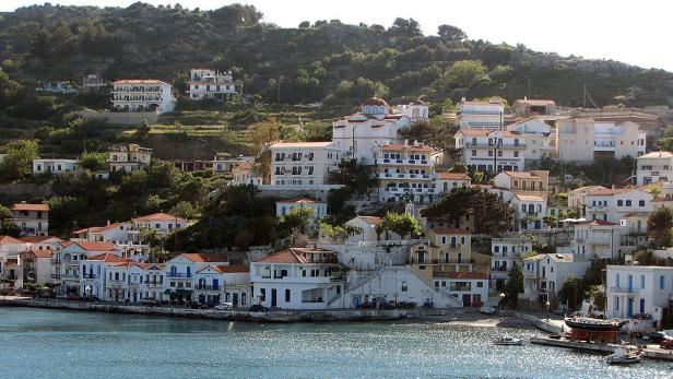 Ein Küstenort mit weißen Häusern und roten Dächern erstreckt sich einen bewaldeten Hügel hinauf.
