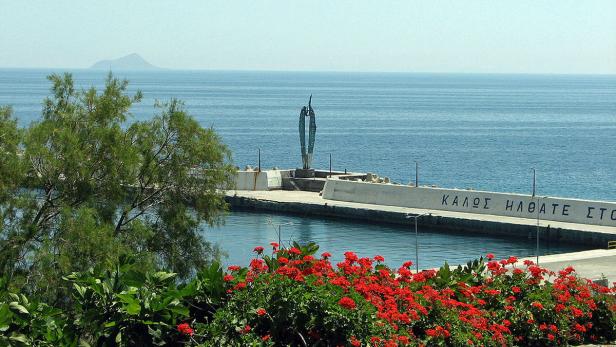 Eine Skulptur steht auf einem Pier mit Blick auf das Meer und eine ferne Insel.