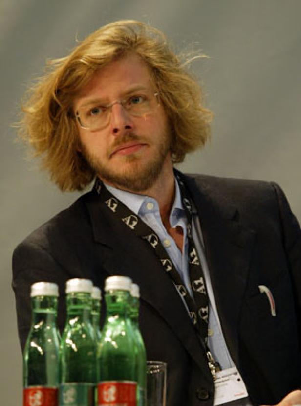 Ein Mann mit blonden, lockigen Haaren und Brille sitzt an einem Tisch mit Wasserflaschen.