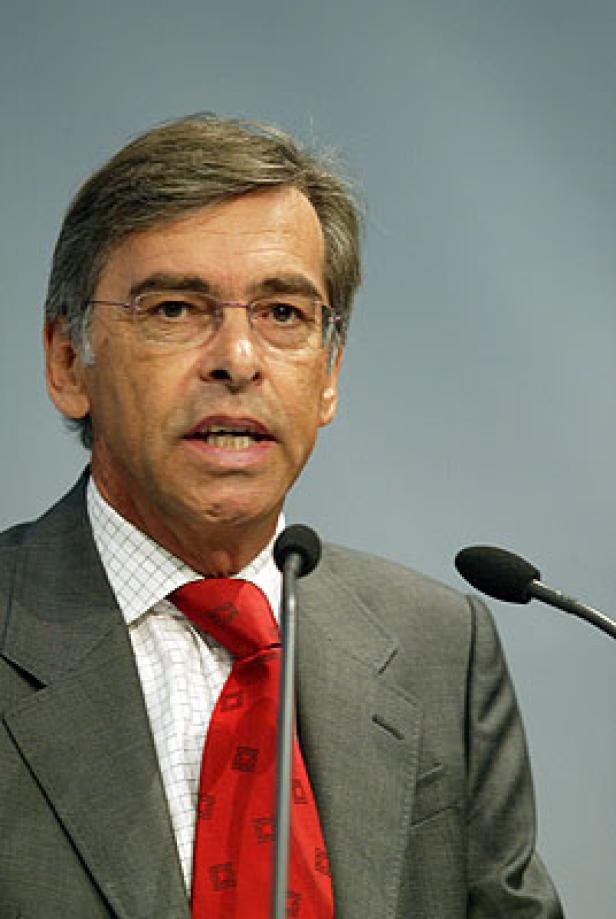 Ein Mann mit Brille und roter Krawatte spricht vor Mikrofonen.