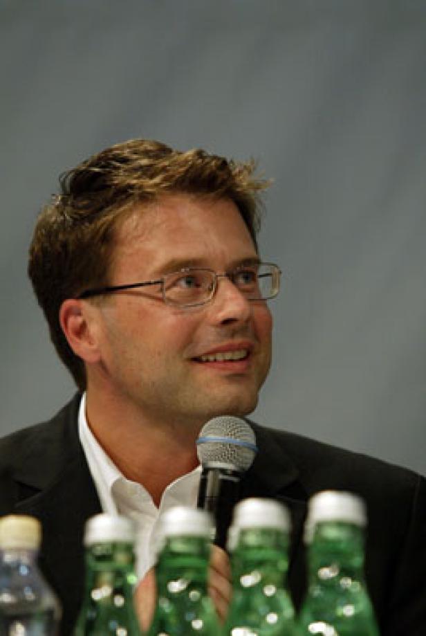 Ein Mann mit Brille spricht in ein Mikrofon vor mehreren Wasserflaschen.
