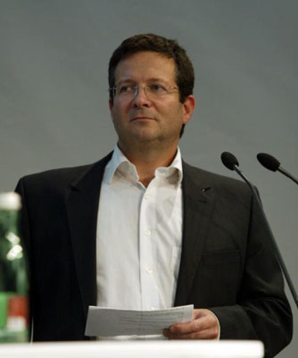 Ein Mann mit Brille hält ein Blatt Papier vor Mikrofonen.