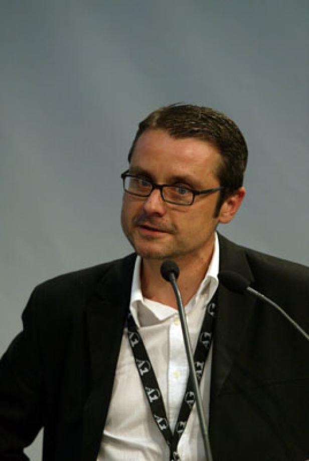 Ein Mann mit Brille spricht bei einer Konferenz in ein Mikrofon.