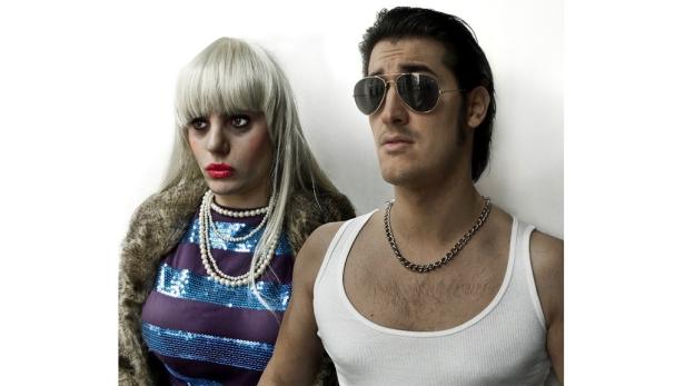 Ein Mann mit Sonnenbrille und eine Frau mit Perlenkette posieren vor einer weißen Wand.