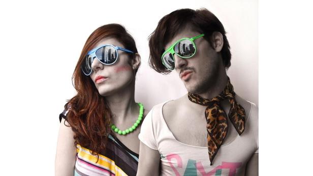 Ein Mann und eine Frau posieren mit farbigen Sonnenbrillen, in denen sich eine Stadt widerspiegelt.