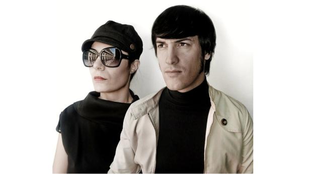 Ein Mann und eine Frau posieren vor einem weißen Hintergrund. Die Frau trägt eine Sonnenbrille, in der sich ein Gebäude spiegelt.