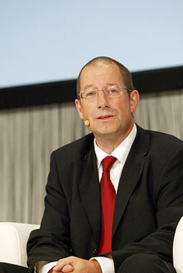 Ein Mann mit Brille, Anzug und roter Krawatte sitzt auf einem weißen Sessel.