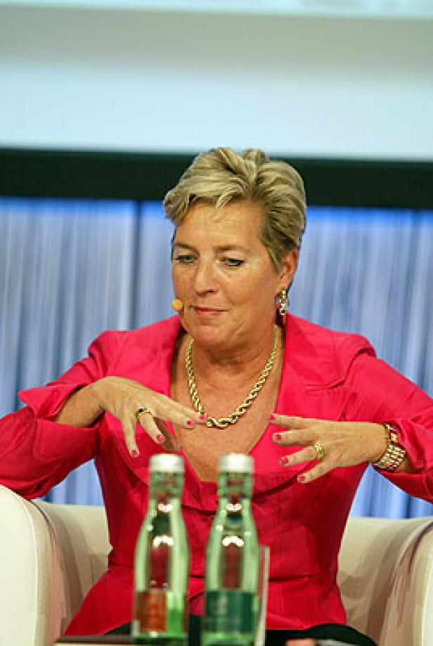 Eine Frau in einem pinkfarbenen Blazer gestikuliert vor zwei Wasserflaschen.