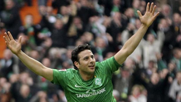 Ein Fußballspieler in grünem Trikot jubelt mit erhobenen Armen vor einer Zuschauermenge.